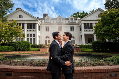Gay couple with arms around each other smiling coming in for a kiss standing in front of the Duke Mansion by Charlotte wedding photographers DeLong Photography