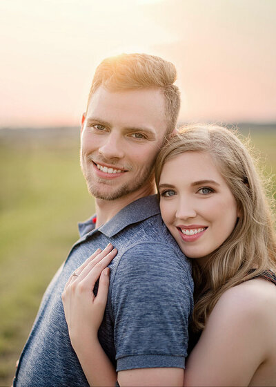 blonde engaged couple hugging and leaning together with sunset sky behind them