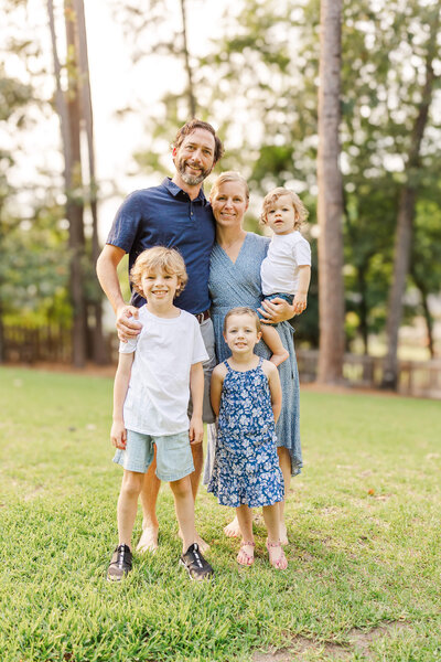 A family of five poses outdoors on the lush grass, encircled by trees. The parents stand behind, with the mother gently holding a young child. In front, two children stand side by side. Casually dressed and smiling at the camera, they capture a joyful moment with their Fayetteville NC family photographer.