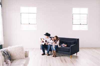 Jackson Hole wedding photographer sits in a white room on a dark couch with her family as they play guitar together
