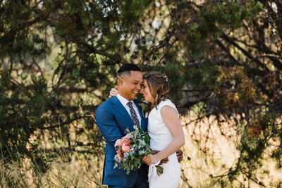 A groom wearing  a blue suit and floral tie laughs with his bride who is wearing a classic high neckline wedding gown outside of their Tetherow Wedding in Bend Oregon. | Erica Swantek Photography