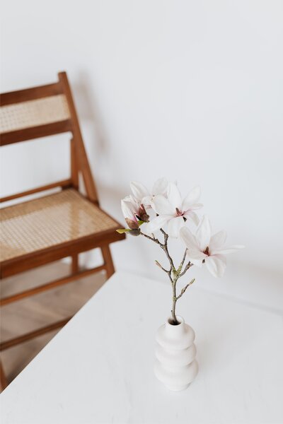 flowers in a vase on top of a white table with a brown chair behind