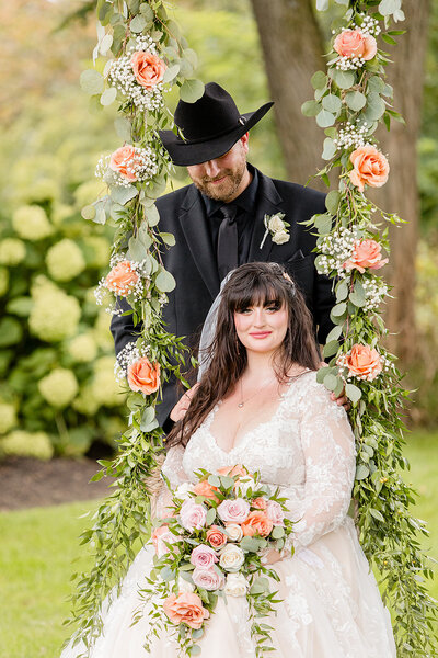bride sitting on floral decorated swing while groom looks down at her lovingly.