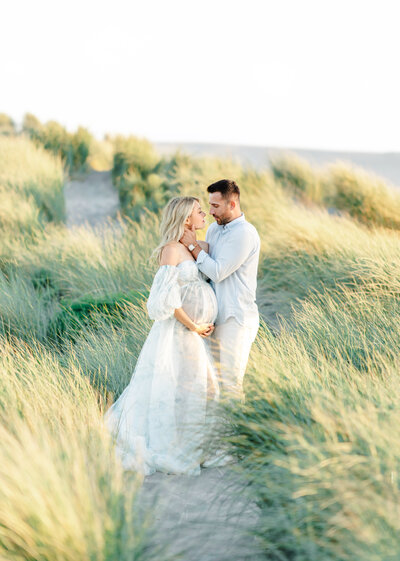 A maternity photography session taken by Bay area photographer shows a mama caressing her baby bump and dad holding her face in a pampas grass beach location.