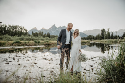 jackson hole photographers photographs bride and groom laughing together as they stand together in the snake river for their grand teton wedding