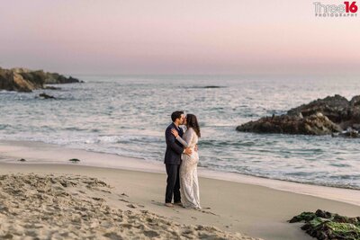 Bride to be looks back at her fiance as he embraces her from behind during a walk on the beach at Woods Cove in Laguna Beach
