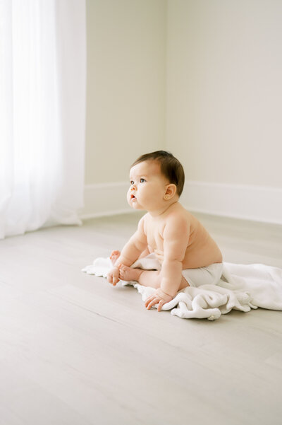 Brunette baby girl sits on white blanket in diaper cover during baby photo session at Raleigh NC studio