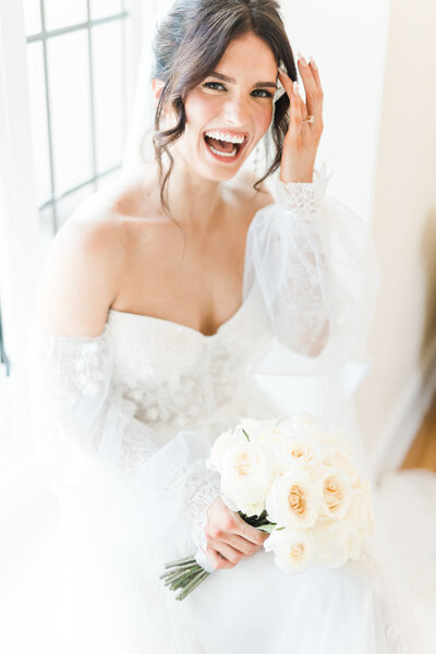 A laughing bride looks into the camera from her perch in a bright window. Her bridal bouquet in her lap, she is having a beautiful day.