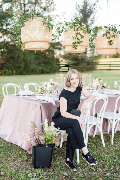 Branding photo for Nashville wedding planner wearing all black and sitting at a styled table with a bucket of flowers on the ground next to her
