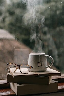 Eyeglasses and steaming mug of tea on a stack of books by a window