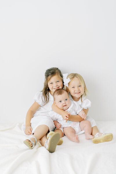 Two little girls holding baby sister, wearing white in a white studio