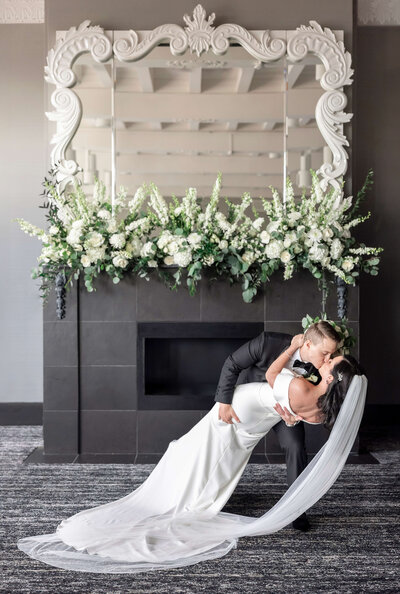 A couple in wedding attire shares a romantic dip-kiss in front of a decorative fireplace adorned with lush white and green floral arrangements. The brides veil cascades elegantly to the floor.