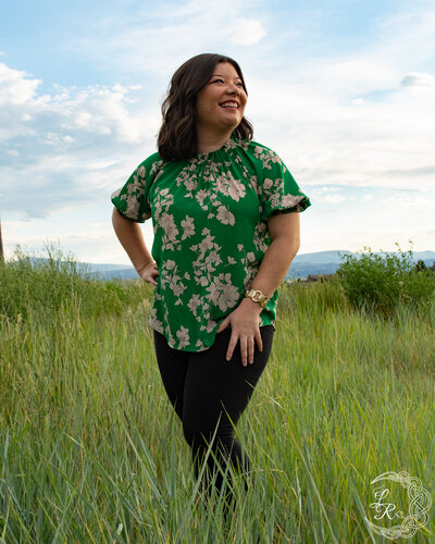 photo of Ashlyn in black pants and a green floral top looking over her left shoulder while standing in a field of grass