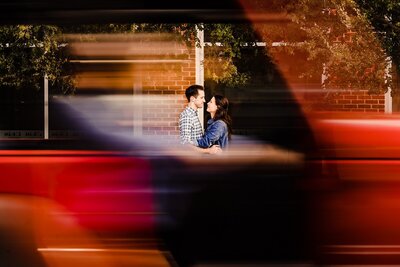 couple embraces on the sidewalk; in the foreground, a red car drives by creating a blurred frame around the couple