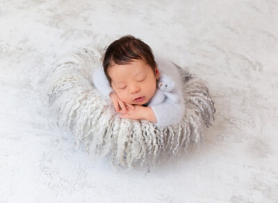 Baby boy is sleeping on his belly wearing a pale blue knit romper with a matching tiny teddy bear tucked under his arm. His hands are folded under his chin. Baby is sleeping atop a white and grey long flokati rug.
