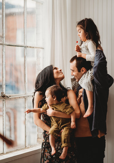 Family of four standing near window of their home. Mom is looking at her daughter on dad's shoulders. Mom is holding baby brother, who is looking out the window.