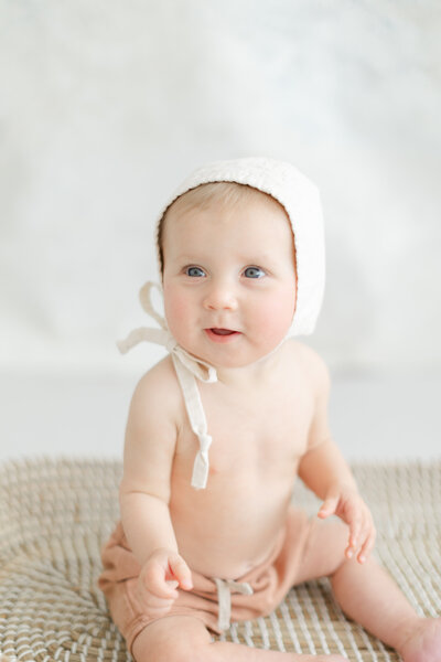 Seated baby smiles while wearing a bonnet