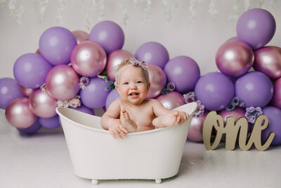 baby girl in white bathtub laughing at camera with her feet on the side of the tub. there are purple balloons in the bakcground
