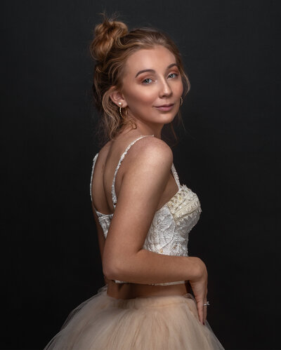 lady in white lace top and cream tulle facing sideways in front of a black background with right hand lightly touching tulle