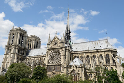 Side view of Notre-Dame Cathedral in Paris, showcasing its Gothic architecture, tall spire, and rose window. The sky is partly cloudy, and trees are visible in the foreground.
