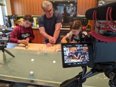 The viewfinder of a camera in the foreground filming a man and two children in a kitchen working on a baking project