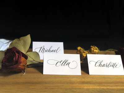 set of three calligraphy place cards against a black background and dried roses intertwined