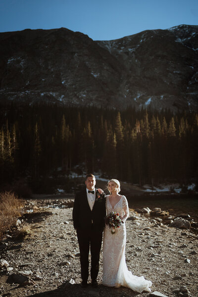 bride and groom are at the base of an alpine lake reading vows to each other. the lake is bright blue, the couple is facing each other and smiling. the bride has a shawl on, and the groom is wearing a tan suit.