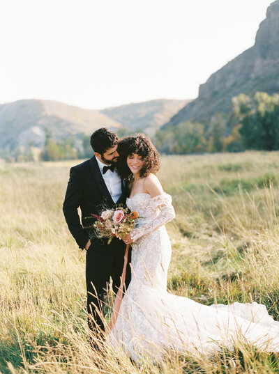 Bride and Groom pose in a field in the mountains, holding a bouquet of flowers