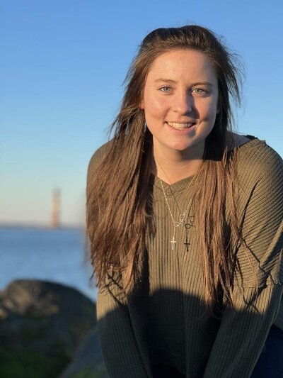 Brunette with long hair and an olive green sweater and a gold cross sitting lakeside smiling at the camera