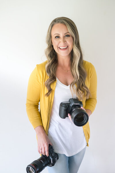 woman smiling holding two cameras wearing a white shirt and yellow cardigan