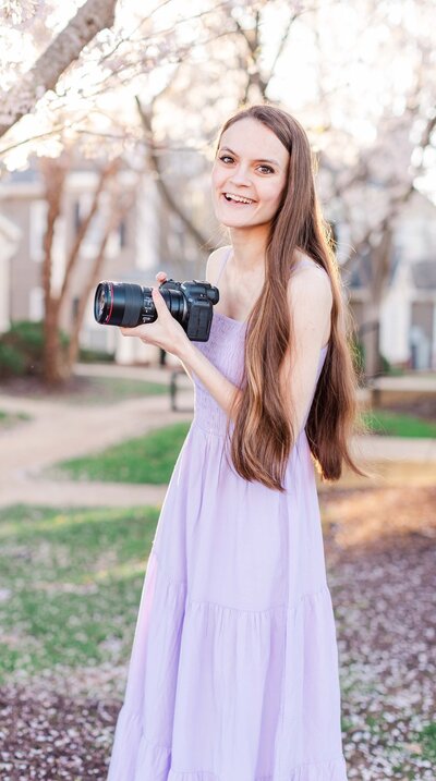 Lady wearing long, lavender dress smiling and holding a camera with cherry blossom trees in the background