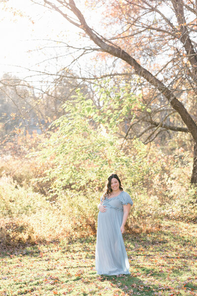 Pregnant woman wearing a light blue dress smiling while cradling her bump in a NJ field in the fall
