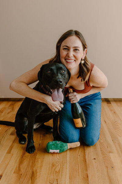 Bridget sitting on her knees on the floor with her arms wrapped around dog, Chloe. Bridget is holding a bottle of champagne and there is a dog toy champagne bottle sitting in front.