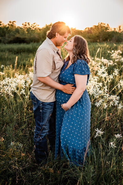 A pregnant couple stand in a field of white flowers and touch noses as mom to be holds her belly.