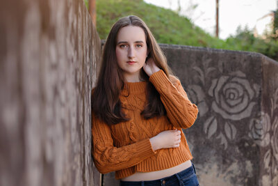Sophisticated Charlotte HS Senior wearing an orange crop sweater and leaning against a concrete wall with stamped rose design