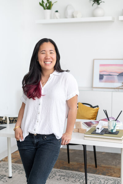 asian woman leaning against her work desk wearing white linen shirt with dark jeans