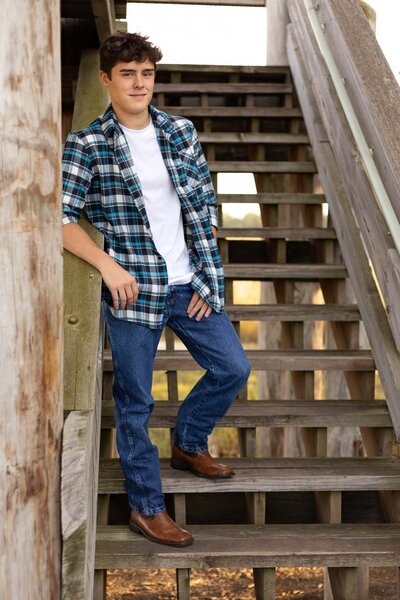 A confident high school senior boy leans against a rustic wooden staircase, dressed in a casual yet stylish plaid shirt over a white t-shirt with blue jeans and brown boots. The natural outdoor lighting highlights his easygoing personality, making this a great example of a laid-back senior photo session with a country-inspired aesthetic.