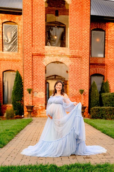 This expectant mom is standing in front of the famous Providence cotton mill she's wearing a Dusty blue dress with one hand cradling her bump and the other holding the train of the dress