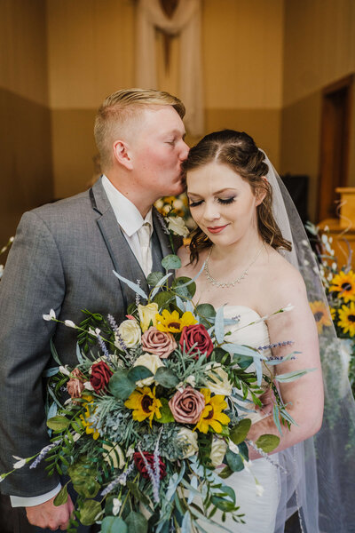 Groom kissing bride's forehead as she holds the bouquet inside a church in nebraska