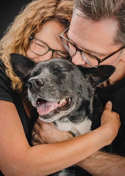 man and woman hugging their dog while smiling with eyes closed