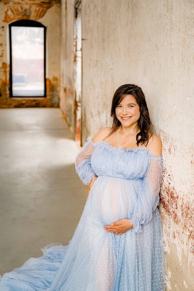 A pregnant woman in a flowing, light blue, off-the-shoulder gown stands against a textured, rustic wall with her hands resting on her belly in a maternity photo