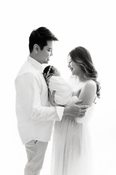 Happy new parents in white stand in a studio cradling their newborn daughter between them