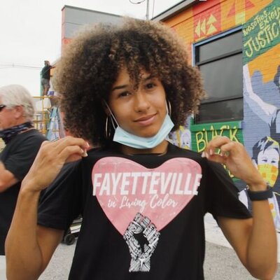 Girl wearing "Fayetteville in Living Color" t-shirt standing in font on black justice mural