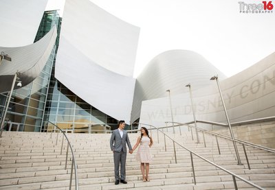 Engaged couple stand on the steps of the Walt Disney Concert Hall holding hands