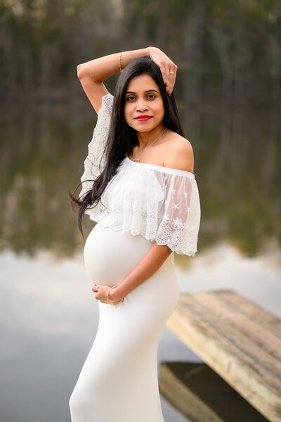 Charlotte Area Mom dressed in a white off shoulder dress standing in front of a lake with one hand cradling her belly and the other arm over her head
