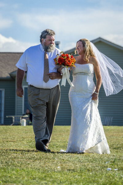 A father walks his daughter down the aisle of a backyard wedding.  The bride is wearing a white wedding dress and her veil is blowing in the breeze.