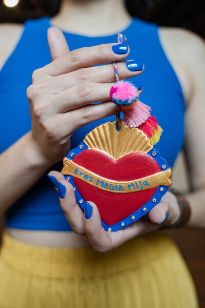 Ursula Rodriguez, founder of Soleil Vida Studio, as she channels creativity and empowerment through her art. In a striking blue top and yellow skirt, adorned with her brand's blue nails, Ursula holds a Mexican heart ablaze with colors – a testament to the vivid experiences awaiting you at our crafting haven.