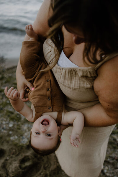 Cute and playful family photography session by Morgan Ashley Lynn Photography on the beach at Lake Michigan in Milwaukee, WI with mom holding baby girl and flipping her sideways, baby girl is laughing
