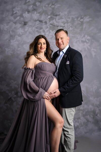 expectant couple in studio session with mom wearing a dusty purple dress and belly exposed and leg out and dad in suit coat and button down shirt and pants