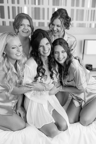 A bride is in a white robe with 5 bridesmaids around her, hugging and smiling towards each other